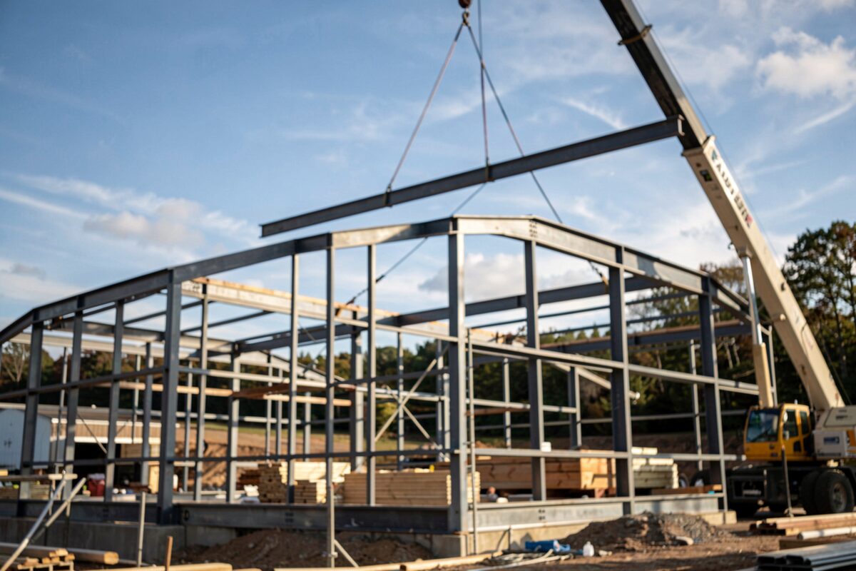 A steel gymnasium frame under construction with a crane lifting a beam against a blue New England sky.
